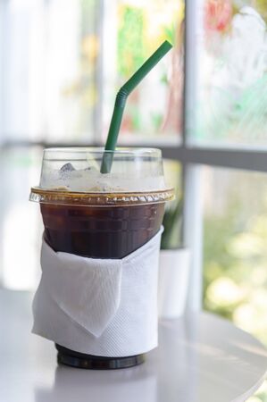 Iced Americano coffee in take home plasttic cup on white table
and window view with green outdoor backgroundの写真素材
