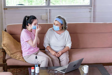 Asian young woman teach older mother, how to wear face mask from online learn in living room with carefully. family conceptの写真素材