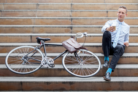 Young businessman sitting on the stairs with bicycle holding paper coffee cup  taking a rest and relaxing  on street in urban cityの写真素材