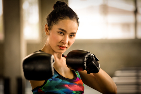 Beautiful athletic Asian Boxer woman punching bag with Boxing Gloves in fitness gym.sport girl in sportswear exercising or workout looking and punch cameraの写真素材
