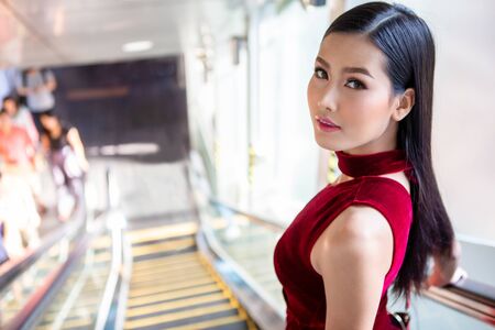 Beautiful young asian woman in red dress going down the escalator in shopping mall . happy elegant lady . copy spaceの写真素材