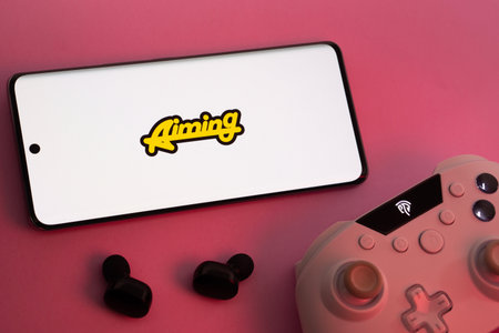 Dhaka, Bangladesh- 08 Apr 2025: A smartphone displaying the Aiming Inc logo next to a gaming controller and earbuds on a pink surface.のeditorial素材
