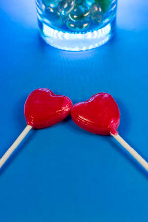Shot of two heart-shaped lollipos next to each other on a blue background with a glass filled with stones.の写真素材