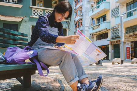 Macau, China - April 05, 2017 - Young girl tourist sitting on the bench in the resting area of A-ma Temple and looking a city map guide to plan a trip in Macau.のeditorial素材