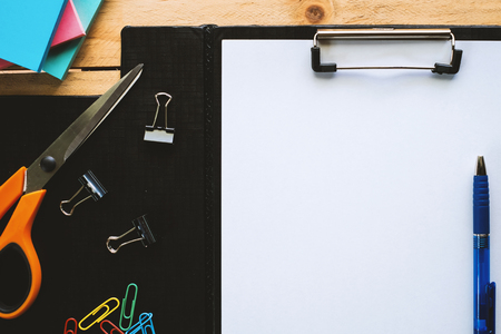 White paper on clipboard with office tool stationery (pen, scissors,notepad, binder and paper clips) on a wooden table backgroundの写真素材