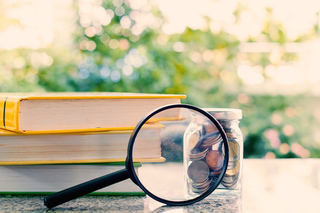 Books and money coins in the glass jar zoomed by the magnifying glass on blurred natural green background and added colour filter for financial and education conceptの写真素材