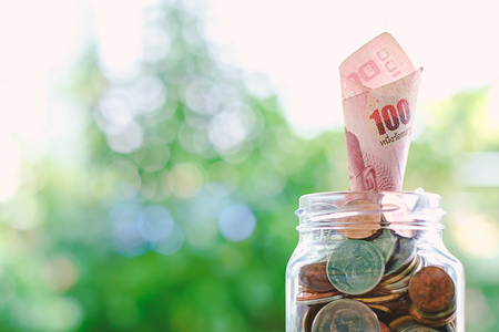 Banknote, 100 Baht Thai currency money growing from the glass jar of coins on blurred green natural...