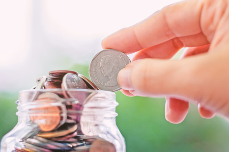 Hand saving a coin into the glass jar on blurred green natural background for business and finance...