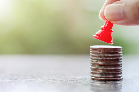 Hand putting a red chess pawn on the stack of coins against blurred natural green background for...