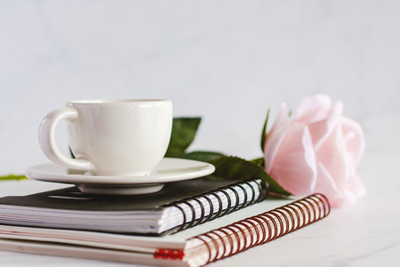 White coffee cup on spiral notebooks with sweet pink rose flower against white marble background for office and school supplies concept, selective focus and DOFの写真素材