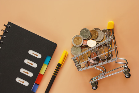 Money and coins in mini shopping cart or trolley with spiral notebook and pen on beige color...
