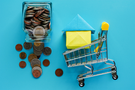 Full of money and coin in the glass jar with shopping cart and house model on blue background for savings, housing and property conceptの写真素材