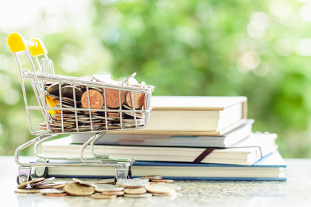 Coins in mini shopping cart or trolley with stack of books against blurred natural green background for save money and education financial conceptの写真素材