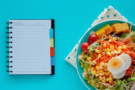 Fresh salad vegetable with boiled chicken egg and blank spiral notebook on blue background for healthy eating and diet food conceptの写真素材