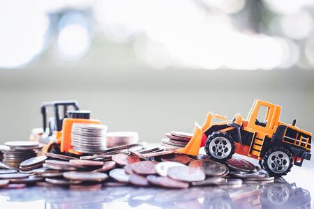 The yellow toy heavy machinery includes bulldozer and forklift with pile of coins against blurred...