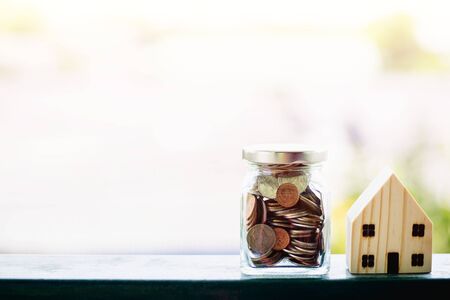 Wooden house model with money coins in the glass jar against blurred natural outdoor background with copy space for business and finance for property conceptの写真素材
