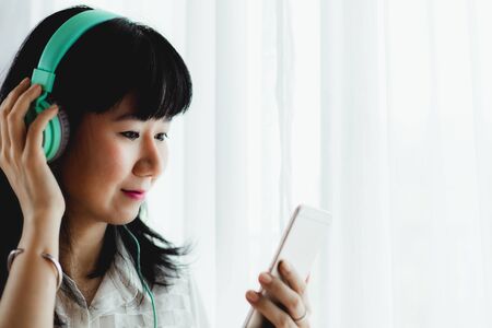 Asian smiling woman wearing headphones, listening music and using smartphone for entertainment and leisure at home concept の写真素材