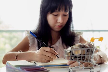Selective focus of Asian woman's hand writing on a notebook with blurred money and coins in a shopping cart for business and financial planning conceptの写真素材