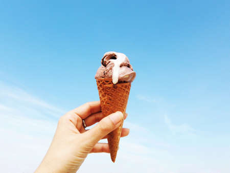 Woman's hand holding wafer cone of chocolate ice cream against bright blue sky background on a sunny...
