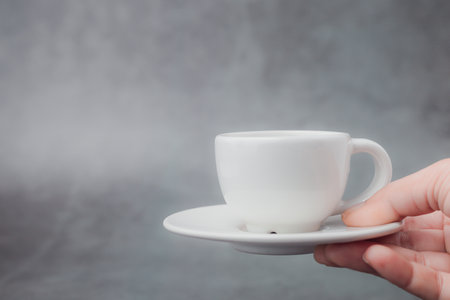 Hand holding white cup of coffee with saucer with copy space on dark background for drinks and...