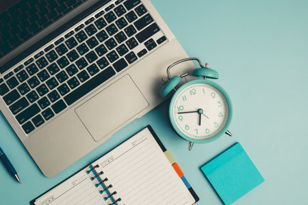 Laptop with an alarm clock, a diary notebook and others work and educational materials on blue background for the concept of work, study and time management.の写真素材