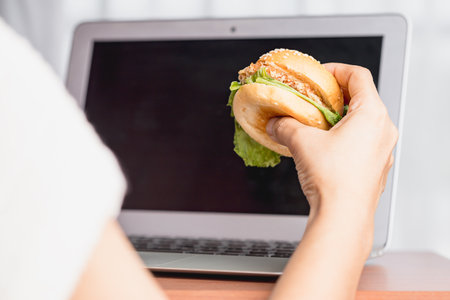 Hand holding a delicious hamburger and using laptop computer for eating while working concept.の写真素材