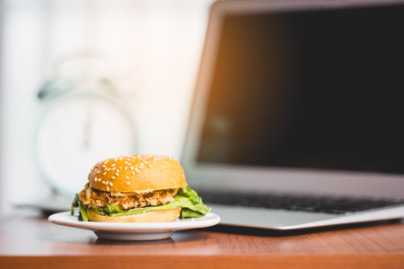 A delicious hamburger on the table with a blurred computer laptop and an alarm clock for eating fast food at work conceptの写真素材