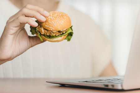 Person enjoying a delicious hamburger meal with cheese, lettuce, tomato, and beef, held in their hands during working for eating fast food at work conceptの写真素材