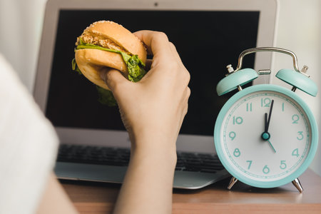 Person eating a hamburger while working, and there is an alarm clock displaying noon time on the work table for the concept of fast food eating at the workplaceの写真素材