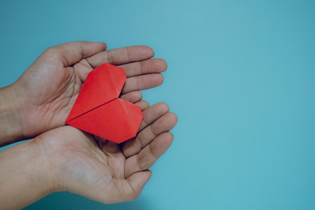 Vibrant red origami heart gently held in cupped hands against a calming blue background with copy...