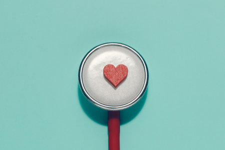 Small red heart rests centrally on a silver stethoscope diaphragm against a blue green background. Symbolizes healthcare, cardiology, heart health, medical care, and compassion in medicine.の写真素材