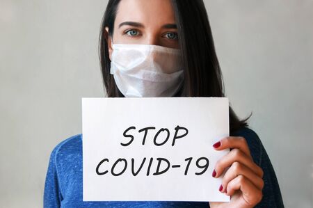 Image of the face of a young woman in a medical mask with a paper in her hand with the inscription STOP COVID-19 .の写真素材