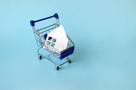 Mini shopping trolley with a white house on a blue background. The concept of buying or renting a house, apartment, cottage and more.の写真素材