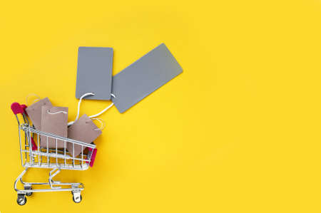 Mini shopping cart with small craft bags and empty gray tags on a yellow background. The concept of shopping in stores and online.の写真素材