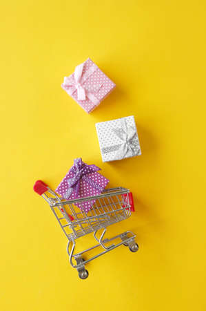 Mini cart with small gifts on a yellow background. The concept of shopping for gifts for the...