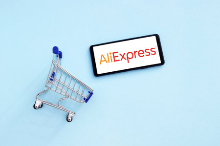 KHARKOV, UKRAINE - NOVEMBER 12,2020: Shopping cart with a smartphone opened AliExpress logo on the screen on a blue background. AliExpress is a global virtual trading platform that provides an opportunity to buy goods from manufacturers from China, as welのeditorial素材