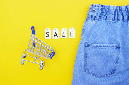 Flat lay, detail of denim jeans, shopping cart with white labels and inscription SALE on yellow background. Shopping and discount concept. Black Friday.の写真素材