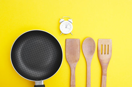 A non-stick skillet, a set of bamboo crockery and a white vintage clock on a bright yellow background. Cooking concept.の写真素材