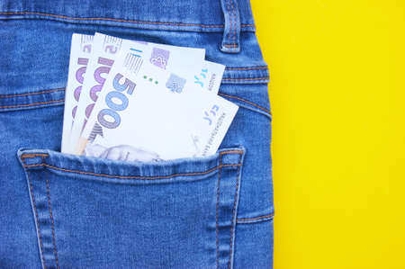 Flat lay, detail of denim jeans with Ukrainian hryvnia bills in pocket on yellow background.