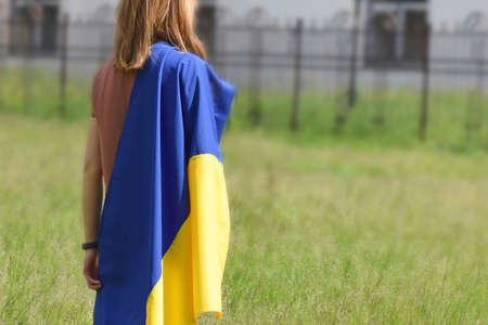 A young girl stands, covered with the national flag of Ukraine, against the backdrop of a local park.の写真素材