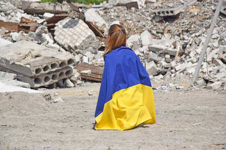 A young girl is on her knees and covered with the national flag of Ukraine against the backdrop of a bombed-out school.の写真素材