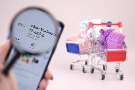 Shopping cart with a smartphone opened Ebay app on the screen on a pink background.