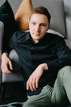 Handsome young man sitting on the floor near the sofa at homeの写真素材