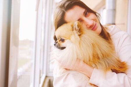 Smiling woman hugging a cute fluffy pomeranian spitz dog looking out the windowの写真素材