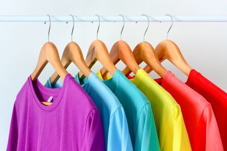 close up collection of colorful rainbow t-shirts hanging on wooden clothes hanger in closet or clothing rack over white backgroundの写真素材