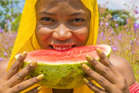 pretty young african lady enjoying a delicious slice of watermelon fruit outsideの写真素材