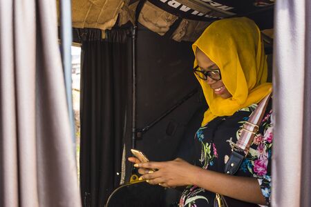 young african woman smiling while using her mobile phone, sitting in the back seat of an auto...