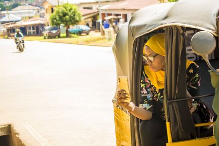 young african woman smiling while using her mobile phone, sitting in the back seat of an auto rickshaw taxiの写真素材