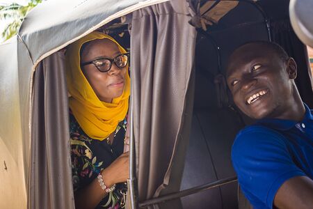 cheerful african man driving a auto rickshaw taxi smiling while listening to a female passenger...