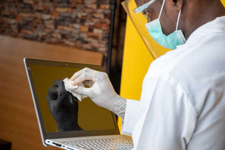 young african man wearing a lab coat and face mask and shield cleaning his laptop computer surface